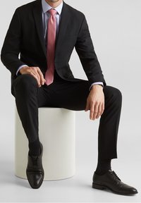 Czarny garnitur o dopasowanym kroju, jasnoniebieska koszula w kratę, różowy krawat, czarne buty eleganckie i czarne skarpetki. Temat siedzi na białym cylindrycznym piedestale.