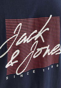 Granatowa koszulka z logo "Jack & Jones" w odważnym białym skrypcie na czerwonym paskowanym tle z napisem "SINCE 1990" poniżej w białym kolorze.