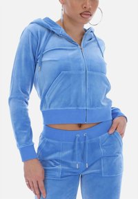 Blue velvet tracksuit featuring a cropped zip-up hoodie with front pockets and matching high-waisted pants with drawstring and pocket accents.