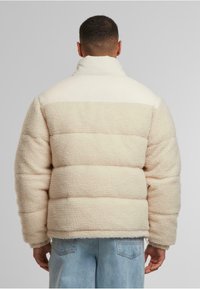 En jakke med puffer i kremfarge med et teksturert, fluffy stoff. Har høy krage, ribbede mansjetter og horisontale quiltede seksjoner.