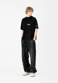 Camiseta negra de gran tamaño con bolsillo frontal y logotipo "MWM" en blanco, combinada con pantalones de denim negros sueltos con rayas laterales y cintura con cordón.