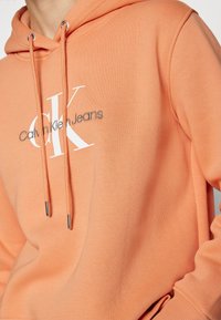 Orange hoodie made of soft fabric, featuring a drawstring hood, front pocket, and white "Calvin Klein Jeans" logo prominently displayed.