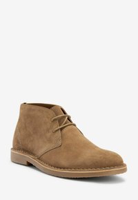 Bota de tobillo de ante marrón con punta redonda, diseño de cordones y suela de goma marrón claro. Presenta costuras mínimas y una textura suave.