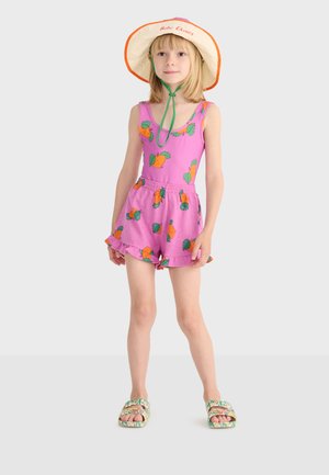 Jeune fille portant une combinaison rose avec des imprimés de fruits orange, un chapeau à large bord avec une lanière sous le menton, et des sandales à motif floral, debout sur un fond gris.