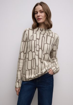 Beiger Pullover mit einem geometrischen braunen Muster, Rundhalsausschnitt, langen Ärmeln und weicher Textur, kombiniert mit dunkelblauen Jeans.