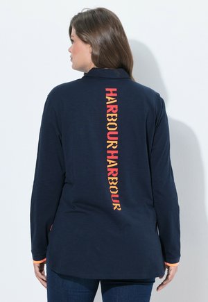 Vrouw met bruin haar die een marineblauw shirt met lange mouwen draagt met het woord "HARBOUR" in rode en oranje letters op de rug.