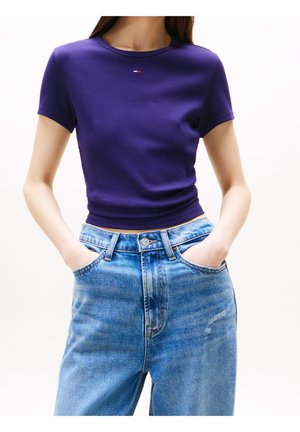 Kobieta w dopasowanym fioletowym krótkim topie z małym logo i jasnoniebieskich dżinsach z wysokim stanem, ręce w kieszeniach.