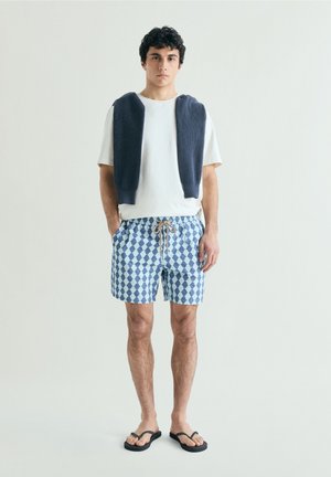 Hombre con camiseta blanca, pantalones cortos azules con estampado, chanclas negras y un suéter oscuro sobre los hombros, sobre un fondo liso.