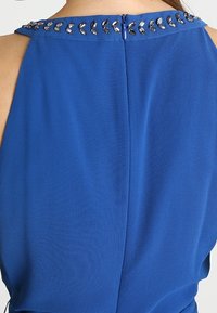 Vue arrière d'un haut sans manches bleu avec une fermeture éclair et des perles argentées décoratives le long de l'encolure.