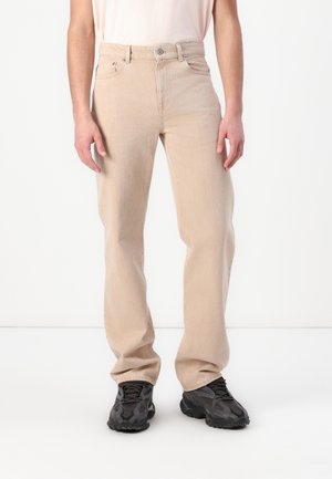 Person, die beige Jeans mit geradem Bein und schwarze Sportschuhe trägt, vor einem weißen Hintergrund stehend.