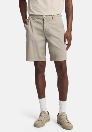 Beige Baumwollshorts mit geradem Schnitt, seitlichen Taschen und Knopfverschluss. Kombiniert mit hellen Sneakers und weißen Socken.