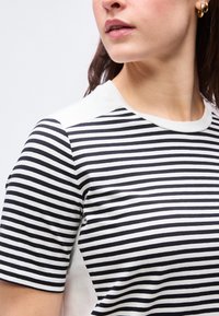 Γυναίκα που φοράει κοντομάνικη μαύρη και άσπρη ριγέ οριζόντια μπλούζα με χρυσούς κρίκους στα αυτιά, εμφανιζόμενη από τους ώμους μέχρι το πηγούνι.