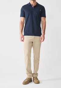 Polo shirt bleu marine avec un col et une poche, associé à un pantalon chino beige et des chaussures en daim marron ; design simple avec un contraste de couleurs subtil.