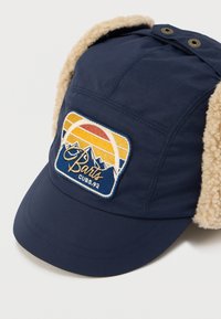 Χειμερινό σκουφί σε ναυτικό μπλε με μπεζ επενδύσεις από φλις στα αυτιά και μια εμπρόσθια επιφάνεια με κέντημα που απεικονίζει βουνά και ηλιοβασίλεμα με το κείμενο «Barts CUBS.92».