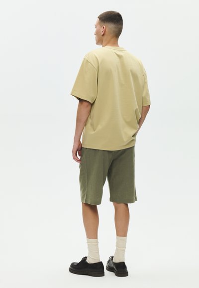 Homme debout, dos à la caméra, portant un t-shirt beige oversize, un short vert olive, des chaussettes crème et des chaussures noires sur fond blanc.