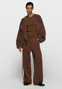 Brauner flauschiger Cardigan über einem braunen Tanktop, kombiniert mit braunen, weit geschnittenen Jogginghosen mit weißem Seitenstreifen und Kordelzugbund.