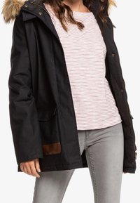 Femme portant un manteau d'hiver noir à capuche avec bordure en fourrure, une chemise à rayures rose clair et blanche, et un jean gris clair.