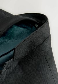 Zwart gestructureerde col van een blazer met klittenbandsluiting en donker patroon aan de binnenvoering op een lichte achtergrond.