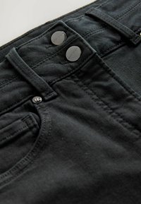 Jeans de mezclilla negra con dos cierres metálicos, presillas para cinturón y detalles de costura prominentes. Tejido texturizado y diseño limpio.