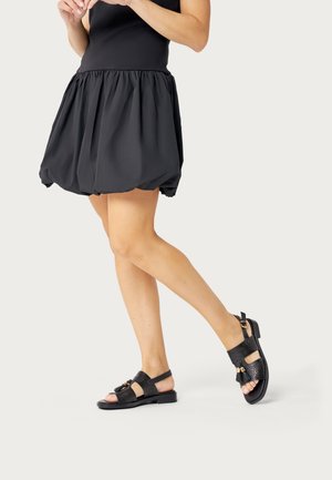 Donna con una minigonna nera arricciata e sandali neri in pelle con punta aperta e dettagli con nappine, in piedi su uno sfondo chiaro.