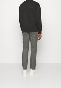 Muž stojící zády, oblečený v černé mikině, šedých kalhotách, bílých ponožkách a bílých teniskách na jednobarevném bílém pozadí.