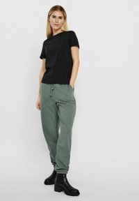 Schwarzes Kurzarmhemd kombiniert mit grünen Jogginghosen, die einen elastischen Taillenbund und Bündchen an den Knöcheln haben, abgerundet mit schwarzen Ankle Boots.