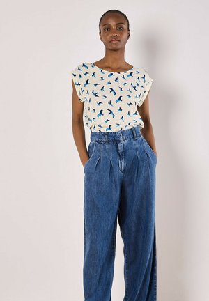 Blouse blanche avec imprimés d'oiseaux bleus, manches courtes, associée à un pantalon en denim bleu taille haute et à jambes larges, avec des plis à l'avant.