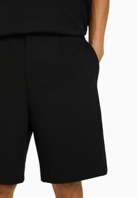 Muž v černých šortkách ke kolenům s rukou v kapse na bílém pozadí.