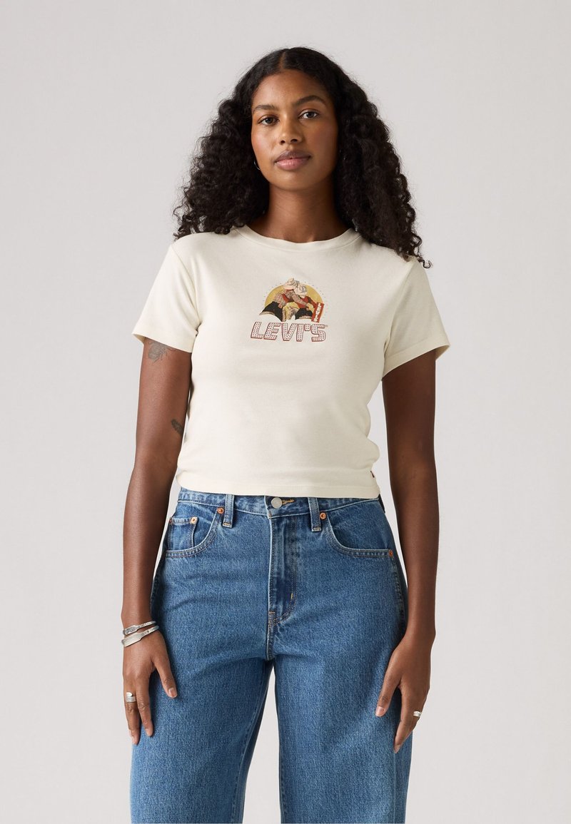 Chemise crème à manches courtes avec un logo graphique "Levi’s" en lettres stylisées. Associée à un jean en denim bleu taille haute.