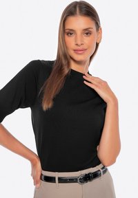 Černý pletený top s krátkými rukávy a kulatým výstřihem. Model má na sobě béžové kalhoty s lesklým černým opaskem. Hladká textura, těsný střih.