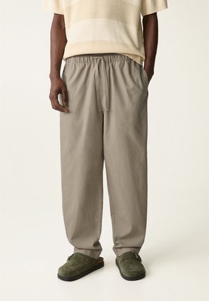 Man wearing beige drawstring pants, light beige short-sleeve knit shirt, and green suede slip-on shoes on a plain background.