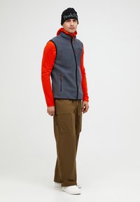 Graue Fleeceweste mit orangefarbenen langen Ärmeln und einer Reißverschlusstasche, kombiniert mit braunen Cargo-Hosen und weißen Schuhen. Schwarze Mütze mit Logo.