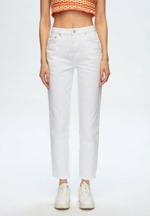 Frau trägt weiße hoch taillierte gerade geschnittene Hose, buntes kurzes Top und weiße Plateausneaker, steht vor einem einfarbigen Hintergrund.