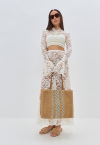 Bijeli čipkasti dvodijelni kroj - cropped top i suknja s razvedenim rukavima, uparen s tkanom slamnatnom torbom sa geometrijskim uzorkom i svijetlosmešim sandalama.