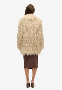Beige fuskpälsjacka med en texturerad, lurvig design. Längd under höften, tillsammans med en brun åtsittande kjol och öppna blockklackskor.