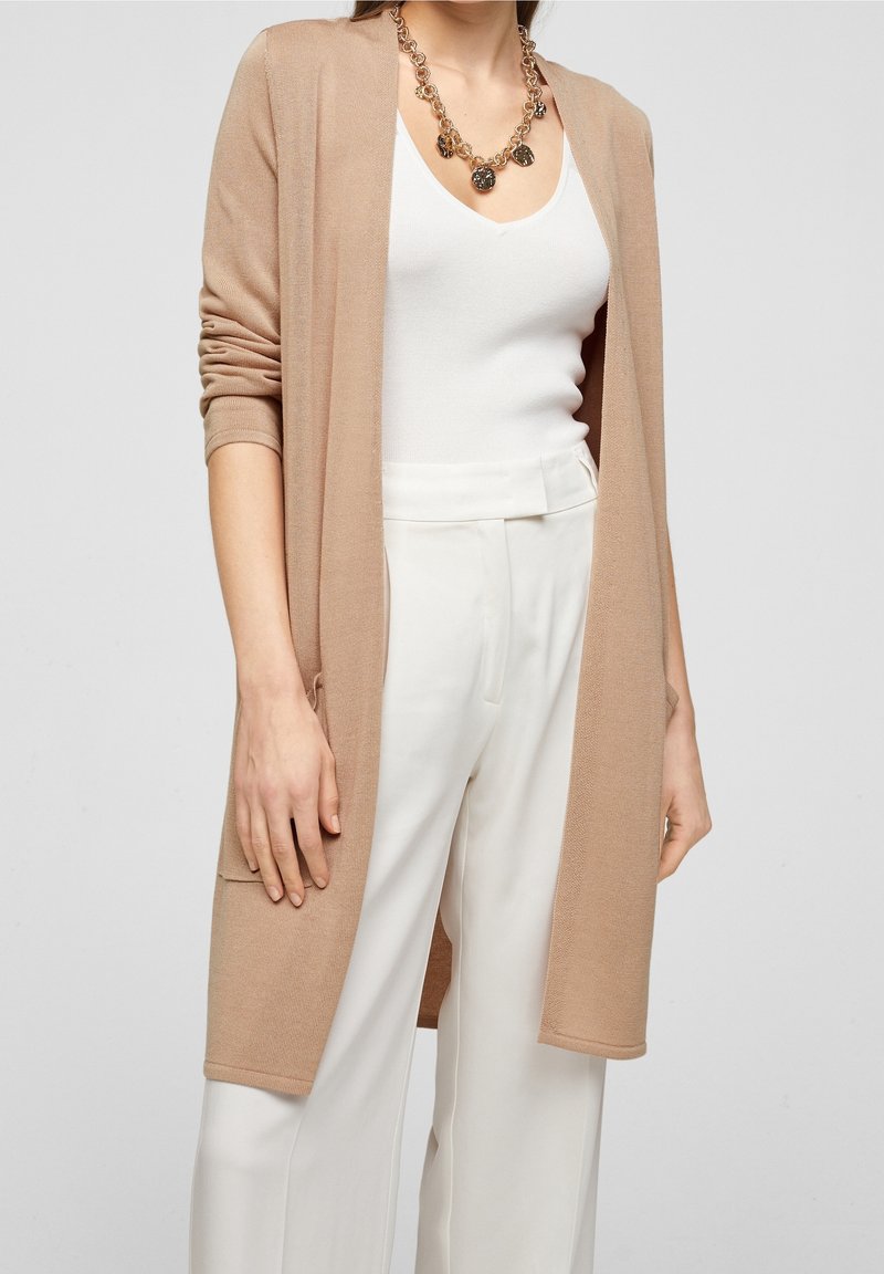 Lätt, beige stickad cardigan med öppen framsida, långa ärmar och sidofickor, bärs över en vit topp och ljusa byxor.