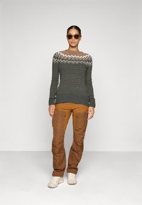 Vrouw in een paarse cargobroek, een gebreide trui met zigzagpatroon, witte sneakers en donkere zonnebril, staand tegen een effen achtergrond.
