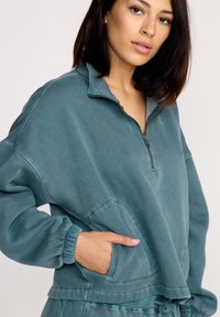 Turkis zip-sweater med høj krave, lange ærmer, ribbestrikkede manchetter og en frontlomme, med en blød, tekstureret stofoverflade.