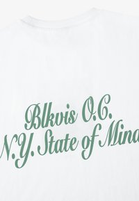 Hvit bomull t-skjorte med grønn tekst som leser "Blkvis O.C. N.Y. State of Mind" i en dekorativ skrifttype på baksiden.