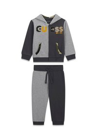 Ensemble de vêtements de détente comprenant un hoodie zippé avec des blocs de couleur gris et noir, le logo "GUESS", et un pantalon tapered assorti avec des poches doublées en camouflage.