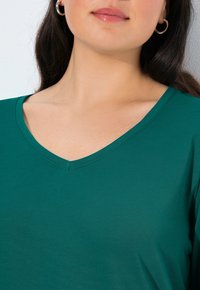 Tyrkysové tričko s výstřihem do V vyrobené z jemného, hladkého materiálu, s uvolněným střihem a čistým prošitím kolem výstřihu a okrajů rukávů.