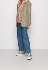 Blazer a cuadros en beige y marrón, combinado con una sudadera rosa. Unos vaqueros anchos azul y zapatillas verde oliva completan el conjunto.