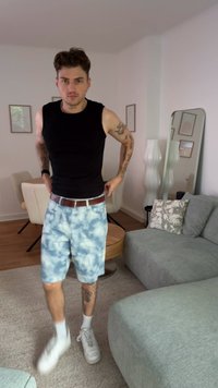 Musta hihaton paita, vaaleansiniset taidevärjätyt shortsit, ruskea vyö, valkoiset tennarit, näkyvät tatuoinnit käsissä ja jaloissa, vaalean värinen asuintila.