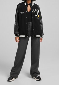 Chaqueta de varsity negra con mangas de cuero, rayas blancas, logotipo bordado de "V", acento floral, combinada con pantalones de pierna ancha grises.
