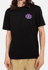 T-shirt en coton noir à manches courtes. Présente un logo rond violet et vert sur la poitrine gauche. Design simple, coupe classique.