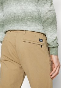Pantaloni beige a gamba dritta, con una tasca posteriore con zip e un piccolo badge con logo. La vita è piatta e dotata di passanti per cintura.