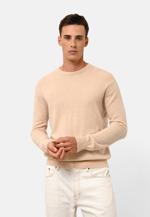Beige knit sweater with a crew neckline, long sleeves, and ribbed cuffs. Soft texture, fitted silhouette, worn over light-colored jeans.