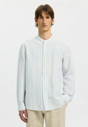 Jeune homme portant une chemise blanche à manches longues avec de fines rayures verticales et un pantalon beige, debout devant un fond clair uni.