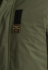 Olijfgroene jacket met een gladde textuur, voorzien van een zwart label met gele tekst. Inclusief een ritszak aan de zijkant.