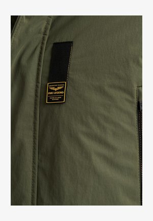 Olivgrüne Jacke mit glatter Textur, versehen mit einem schwarzen Etikett mit gelber Schrift. Enthält eine mit Reißverschluss verschlossene Tasche an der Seite.